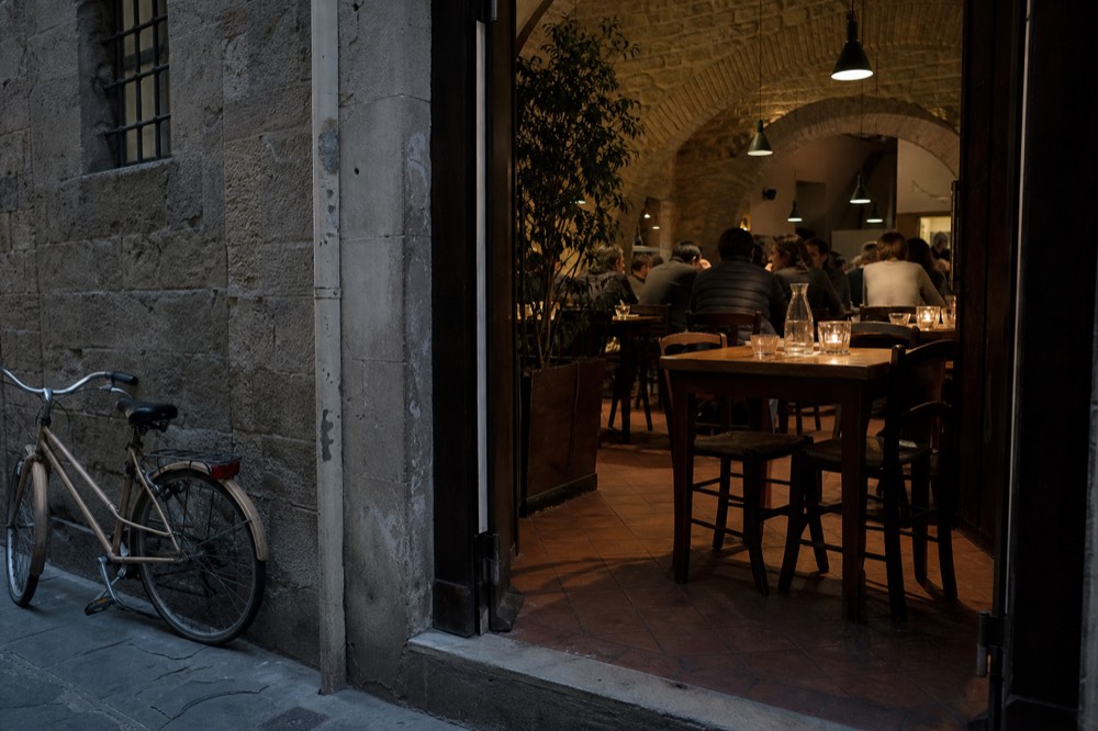 A tiny Florentine osteria interior with closely spaced tables and a chalkboard menu, no text, no logos, no people