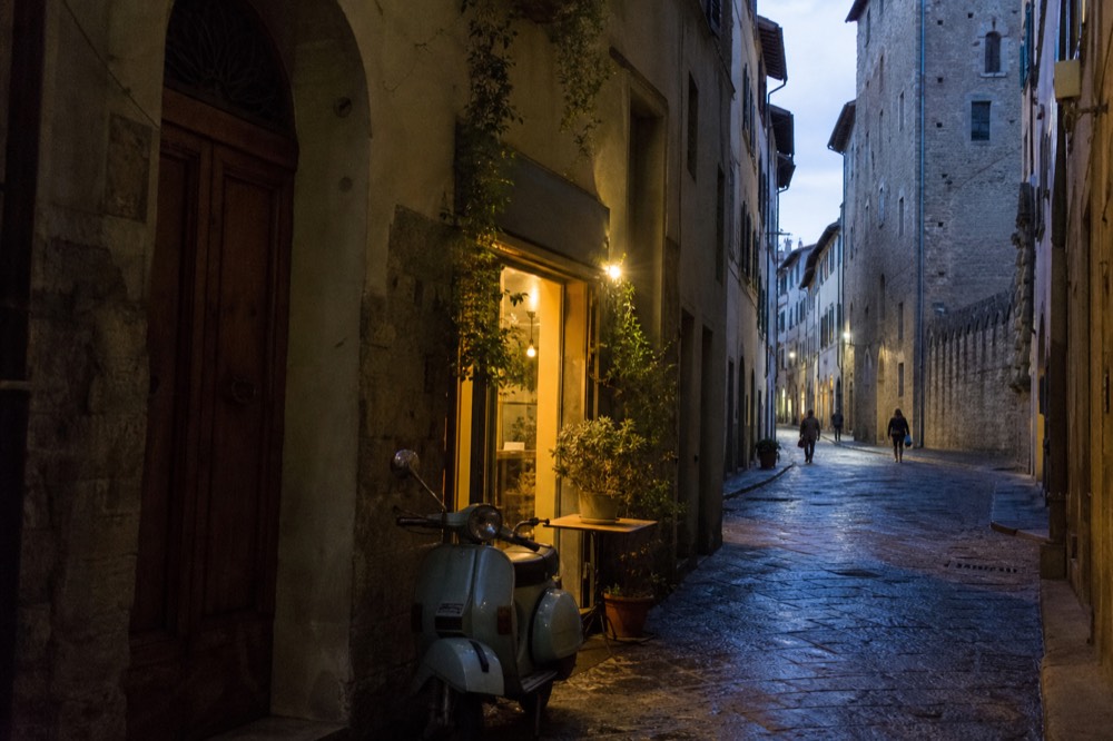 A narrow Oltrarno street at dusk with warm light spilling from a trattoria doorway, no text, no logos, no people