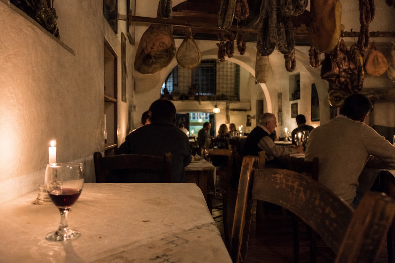 A candlelit Tuscan dining room with rustic wooden tables and hanging cured meats, no text, no logos, no people
