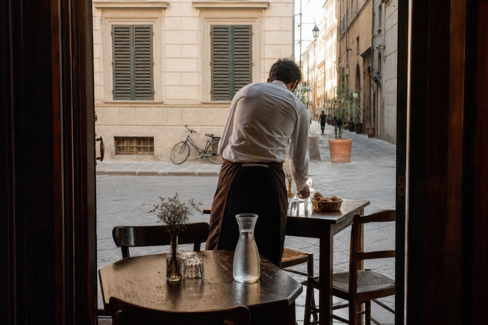 A waiter setting a table with cutlery and a bread basket in a small trattoria, no text, no logos, no people