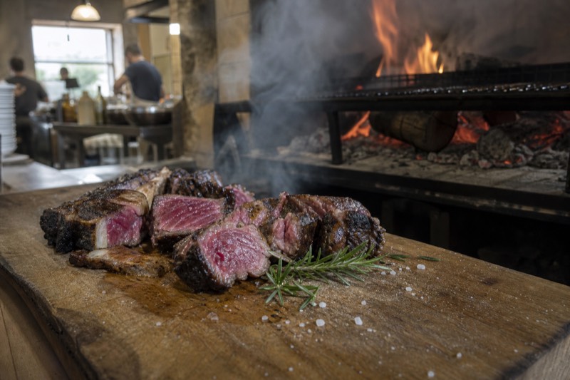 A thick bone-in Florentine steak, crust dark from the grill and interior deep pink, resting on a wooden board with coarse salt, a carving knife and a glass of red wine beside it