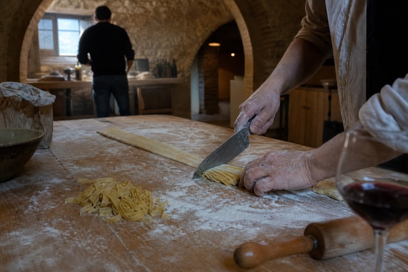Flour-dusted hands rolling fresh egg pasta dough on a wooden board in a Florentine kitchen, a rolling pin, scattered semolina and a glass of red wine waiting at the edge of the counter