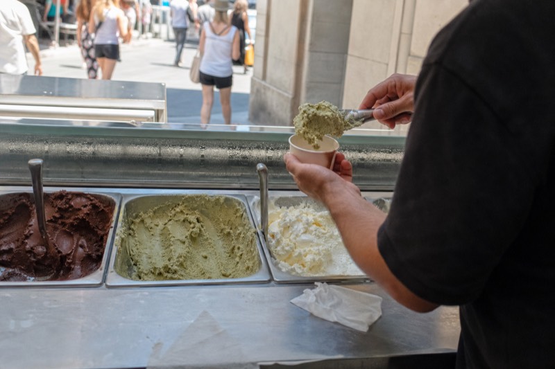 An artisan gelato counter with flat metal tubs of pistachio, dark chocolate and cream gelato, a spatula lifting a pale scoop into a paper cup while a customer waits