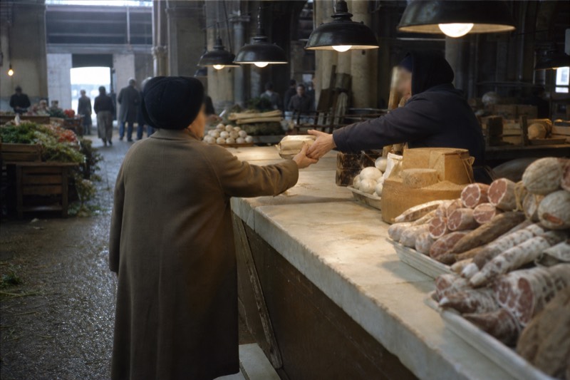 A busy Florentine market hall with stalls of cured meats, wheels of pecorino and fresh produce under warm hanging lights, a vendor handing a tasting across the counter to a small tour group