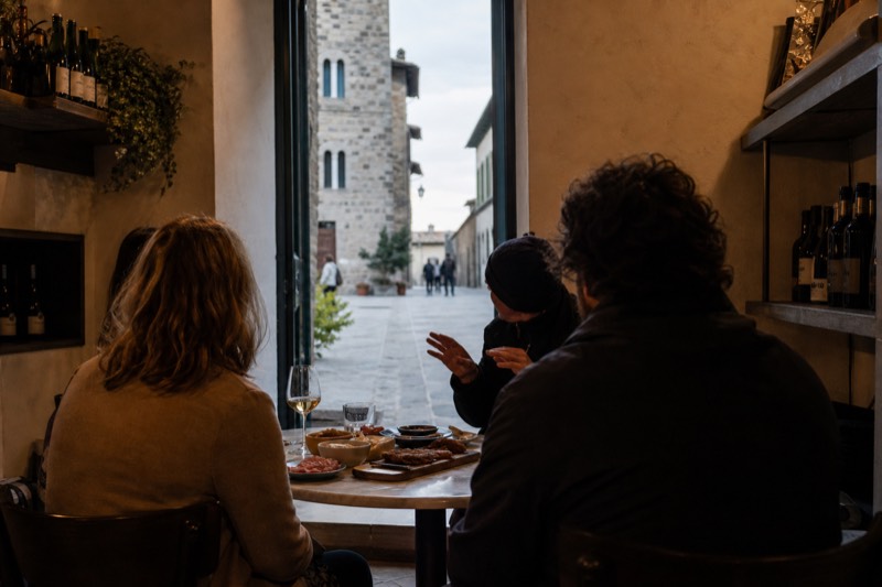 A local guide sharing tasting plates with two travellers at a small marble table in a quiet Florentine enoteca, wine glasses and a cheese board between them, the evening street visible beyond the doorway