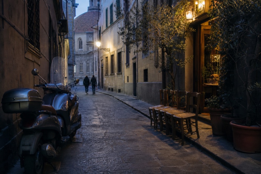 A short queue of empty chairs outside a lit trattoria entrance at dusk, no text, no logos, no people