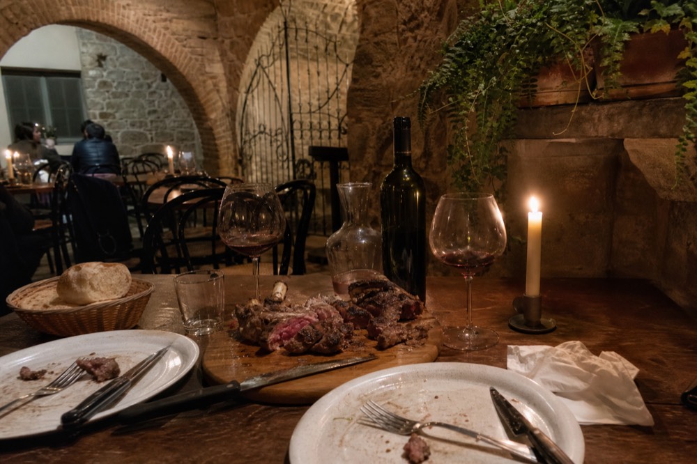 Two place settings sharing a carved bistecca alla fiorentina with a bottle of red wine, no text, no logos, no people