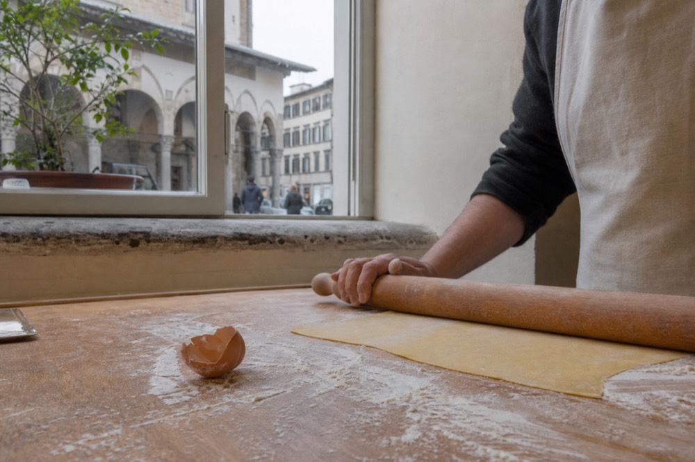 Fresh egg pasta dough being rolled thin by hand with a rolling pin on a wooden board, no text, no logos, no people