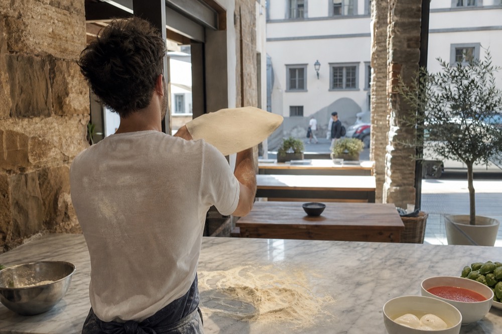 A cook shaping pizza dough by hand on a marble counter with flour and fresh toppings nearby, no text, no logos, no people
