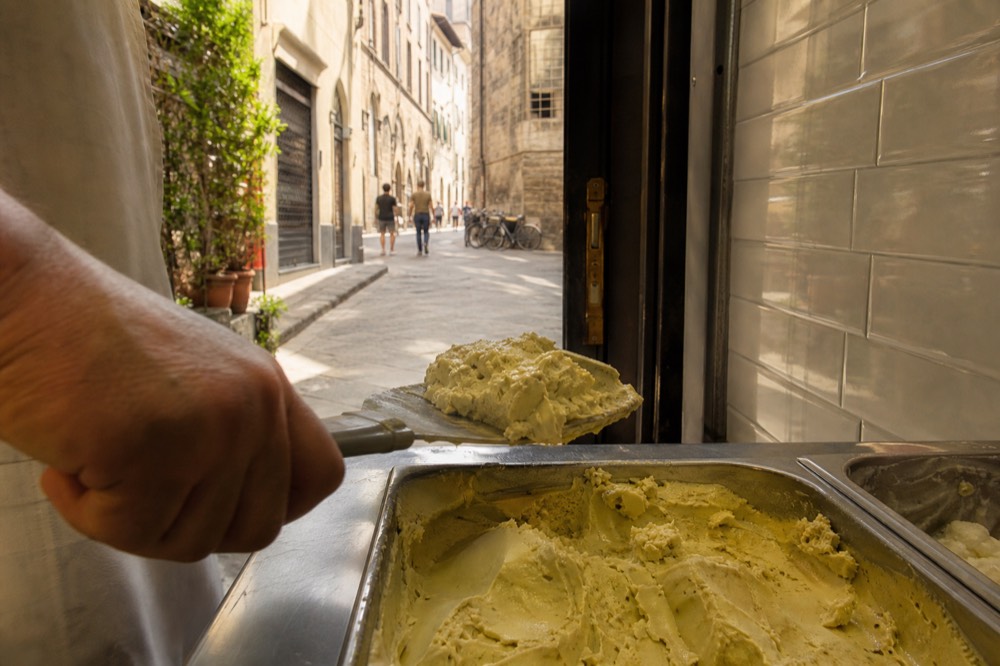 Gelato being scooped from a metal churn pan in a small artisan kitchen, no text, no logos, no people