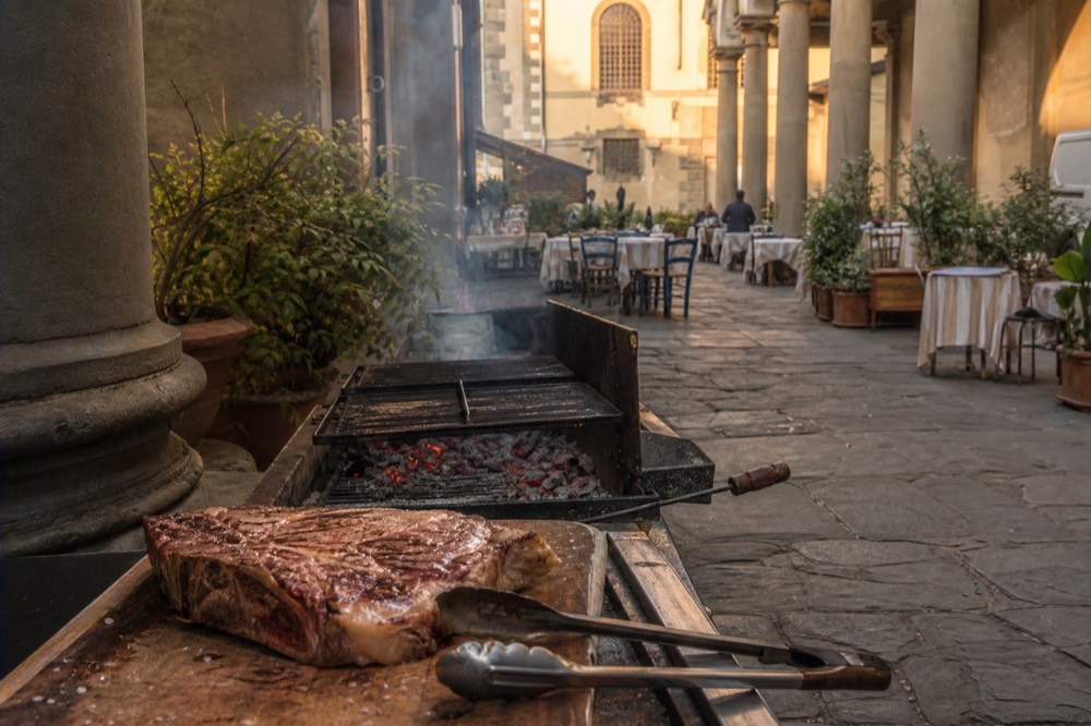 A thick Florentine T-bone steak resting on a wooden board beside charcoal embers, no text, no logos, no people