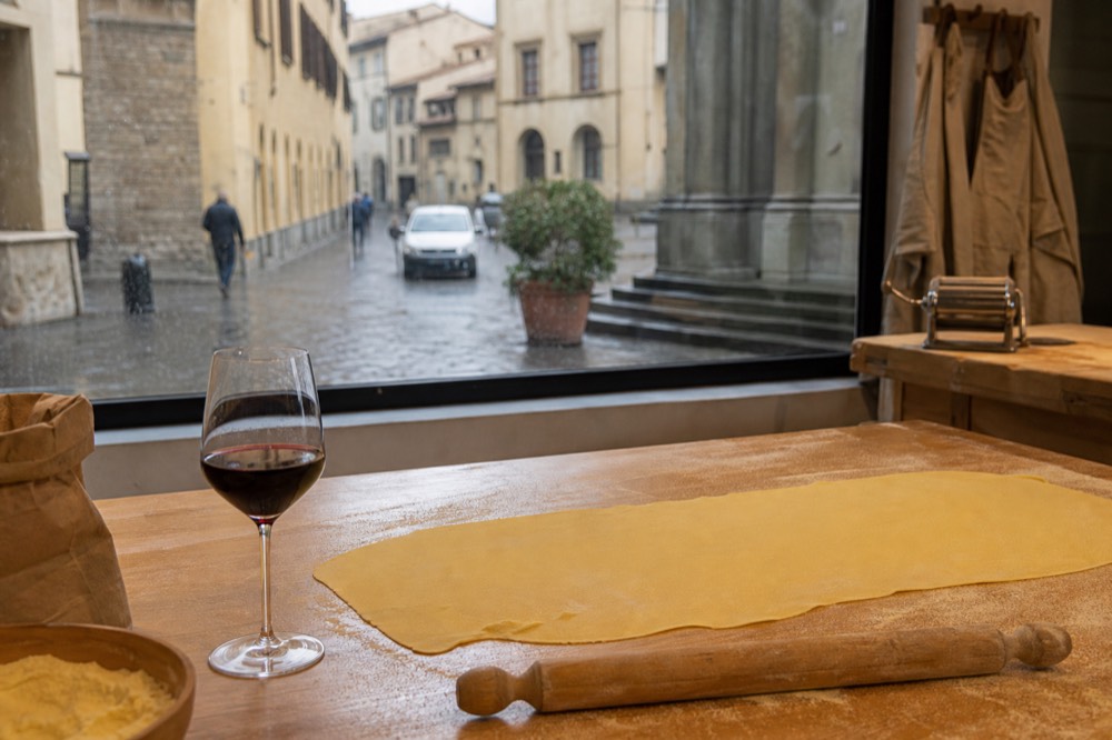 Hands rolling fresh pasta dough on a floured wooden board beside a glass of red wine, no text, no logos, no people