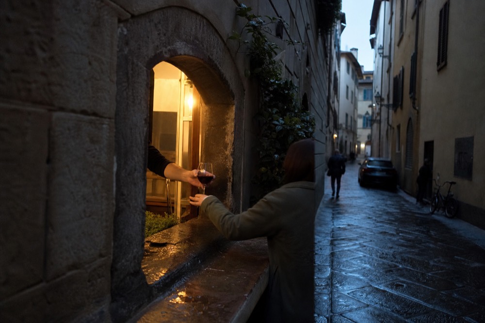 A bartender passing a glass of red wine through a small arched window opening to a customer outside, no text, no logos, no people