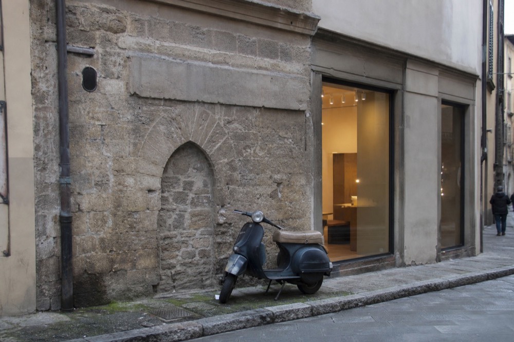 A stone wall with a small filled-in arched opening beside a modern shopfront, no text, no logos, no people