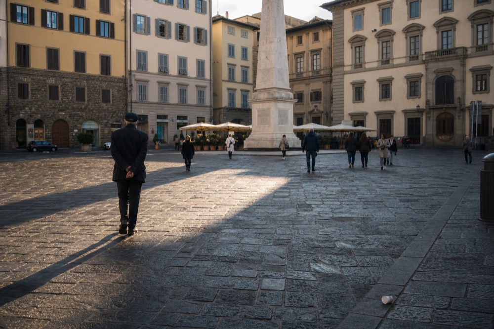 Late afternoon obelisk square in San Lorenzo, tour group shadows on cobbles, shuttered facades, tipped cup, no text, no logos, no people