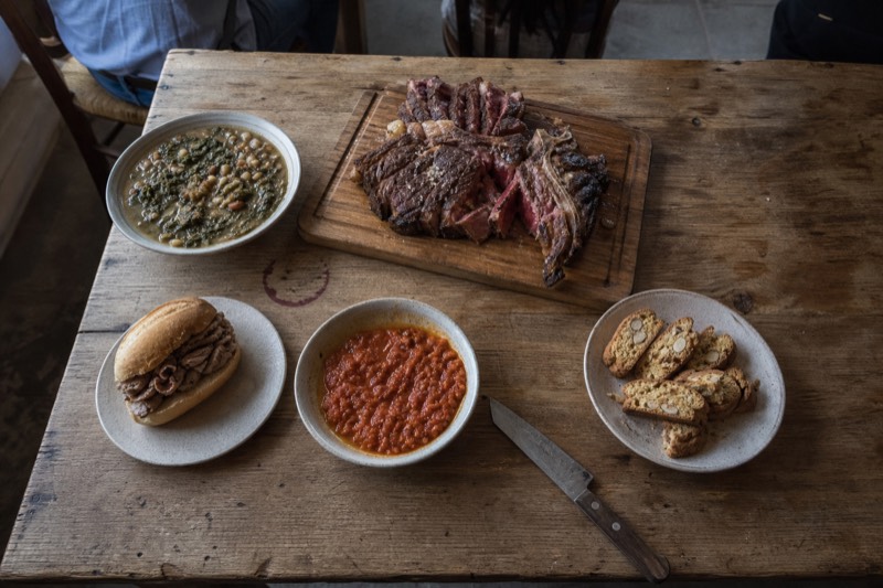 An overhead of the Florentine repertoire on a scrubbed wooden table: a bone-in bistecca on a board, a bowl of ribollita, a lampredotto roll, pappa al pomodoro and a plate of cantucci