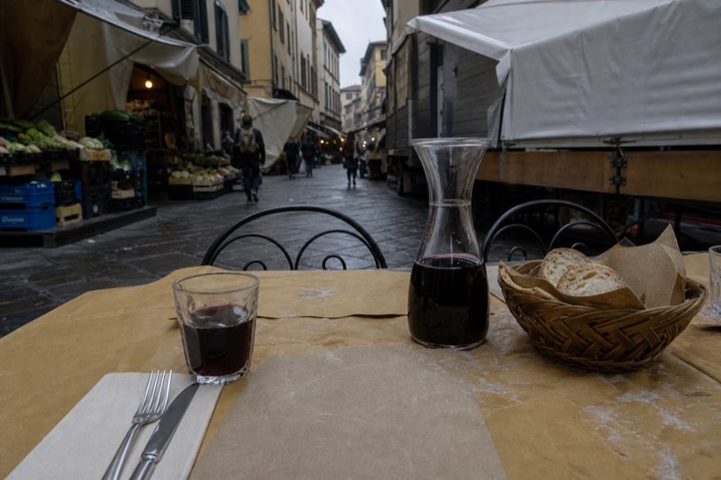 A printed Italian menu on a paper-covered trattoria table showing the per-person coperto line, beside a carafe of house red and a basket of unsalted Tuscan bread