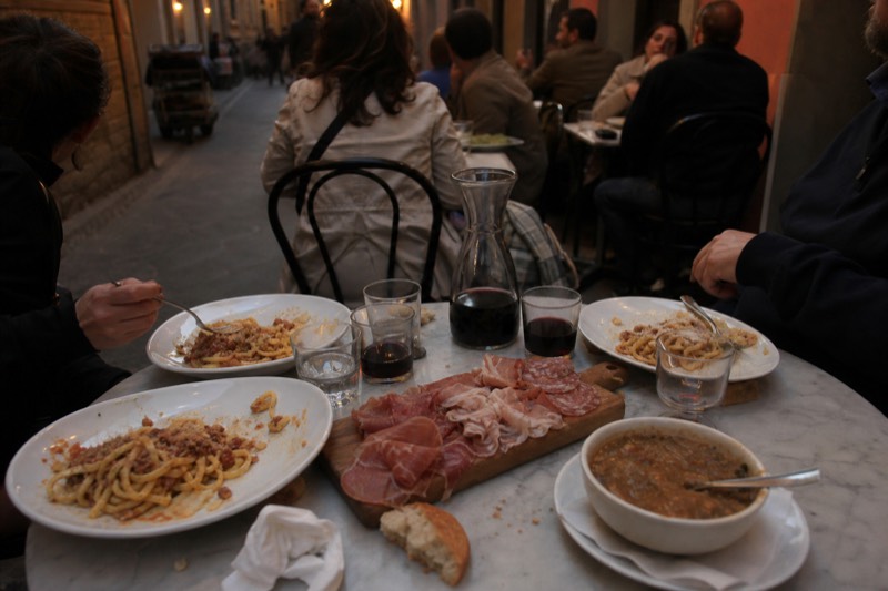 Four people at a shared marble table in Florence deciding between plates of pasta, a bowl of ribollita and a board of cured meats, carafe of red wine between them