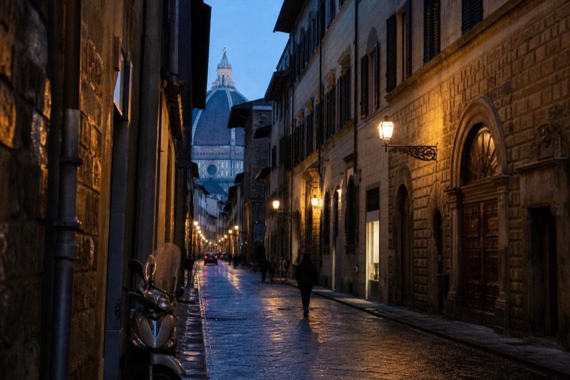 A narrow Florence street of shuttered stone palazzo windows at dusk, the Duomo dome visible at the end of it, warm lamps on in the ground-floor rooms