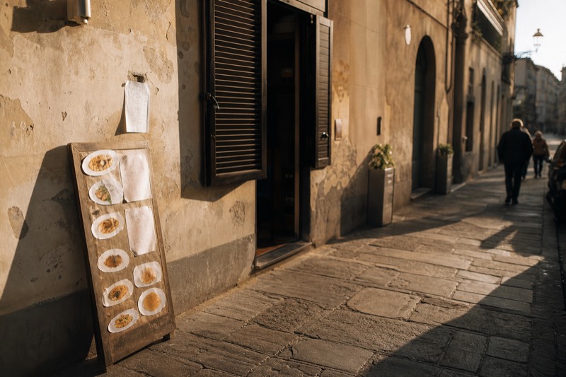 A chalk menu board propped in a Florence trattoria doorway listing the day's dishes and the cover charge, shutters half closed behind it in late afternoon light